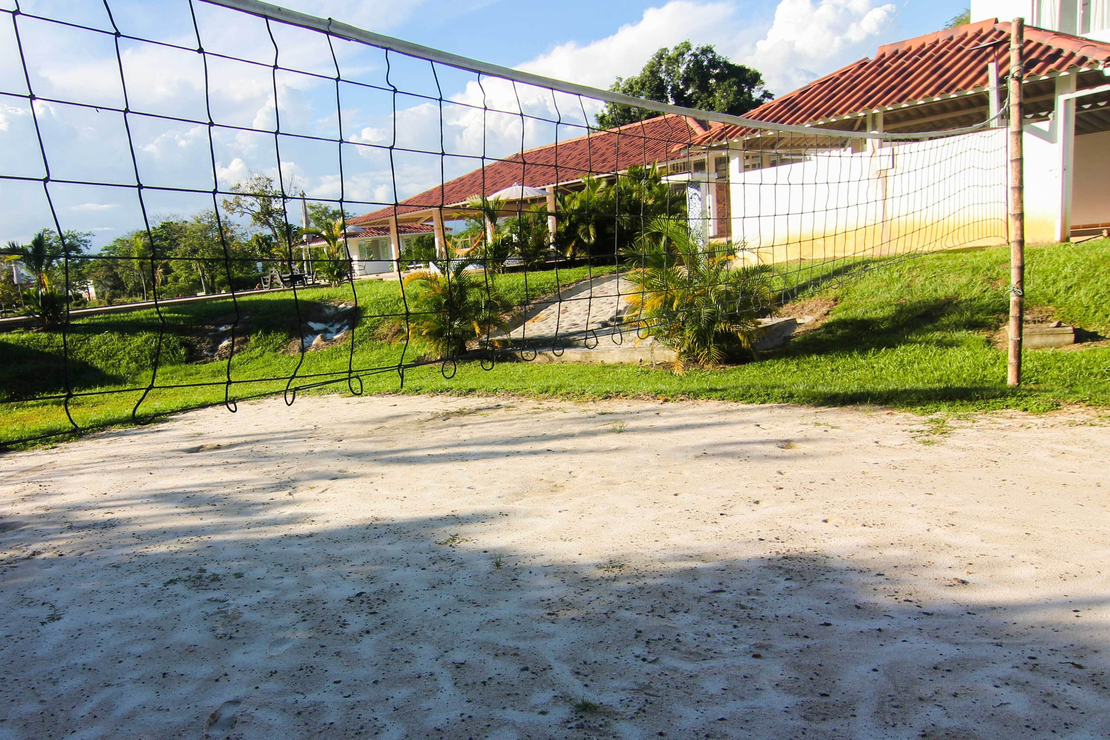 Un terrain de volley-ball avec un filet, entouré d’une zone sablonneuse et, au fond, un bâtiment avec un toit rouge.