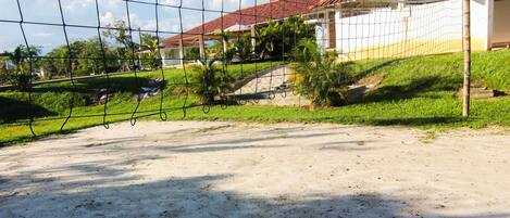 Un terrain de volley-ball avec un filet, entouré d’une zone sablonneuse et, au fond, un bâtiment avec un toit rouge.