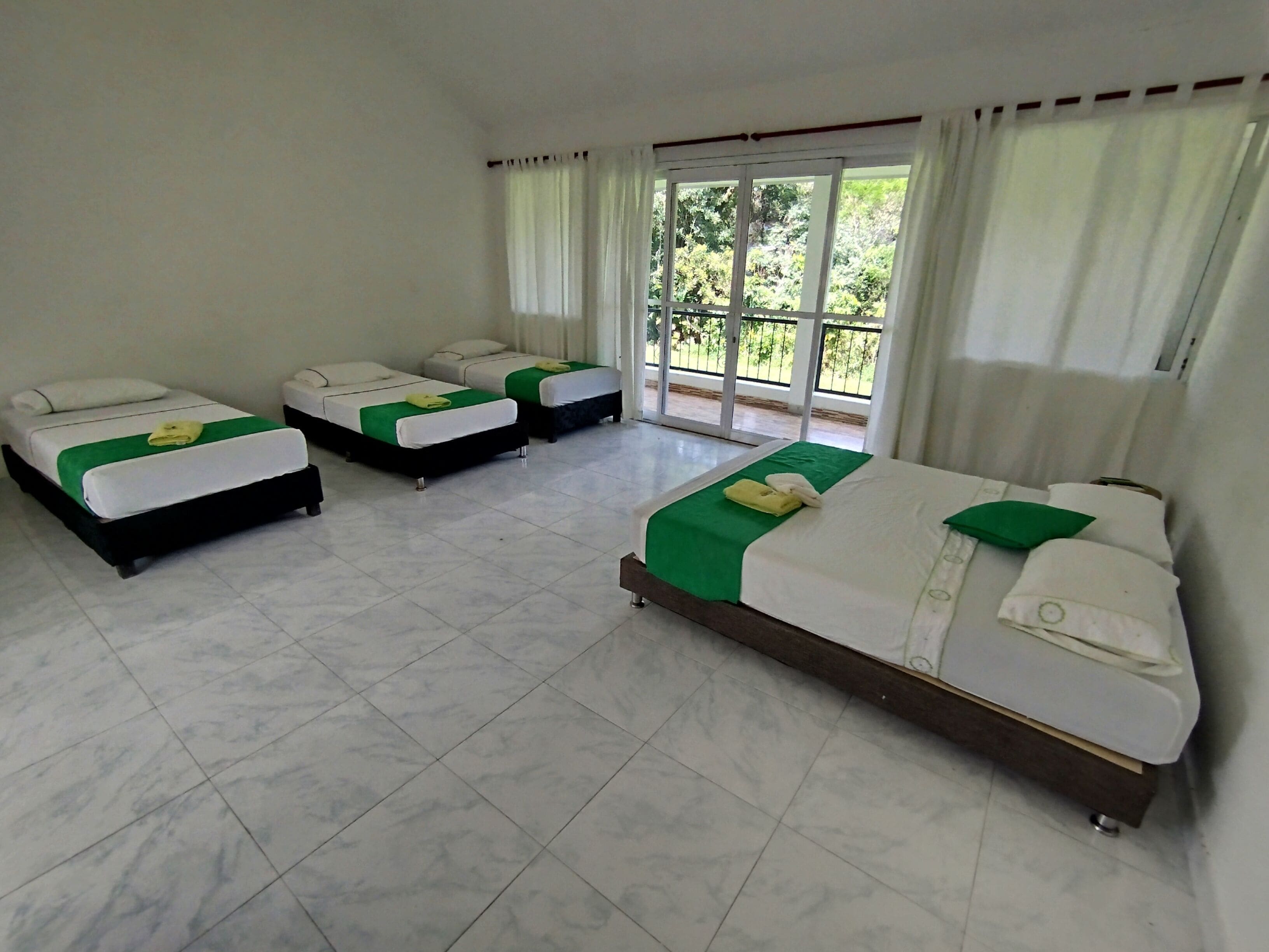 Habitación de hotel con tres camas individuales, cada una con cubrecama verde y almohadas blancas. Las paredes son blancas, el piso está cubierto de baldosas y hay ventanas amplias con cortinas blancas.