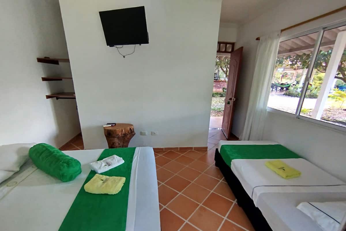 Habitació d'hotel amb dos llits, cadascun amb una colcha verda i roba de llit blanca. Hi ha un petit tamboret de fusta amb un llibre a sobre, un televisor fixat a la paret i una finestra amb cortines.
