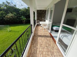 Un balcón con piso de madera, muebles blancos y vista a un jardín verde con árboles.
