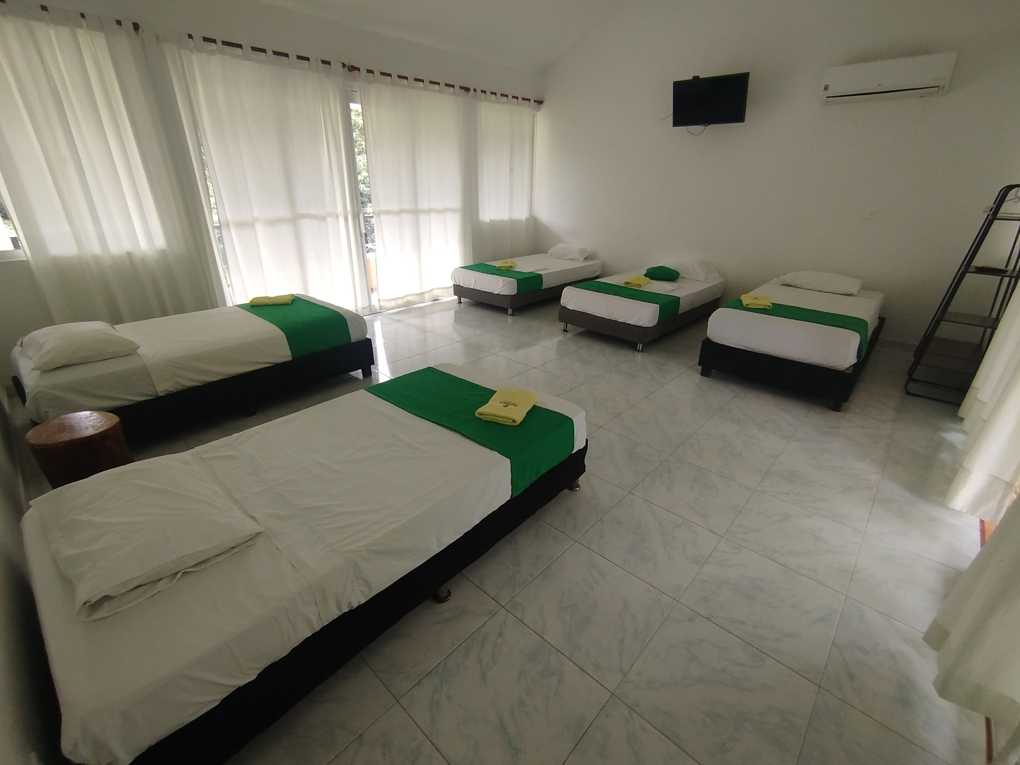 Une chambre d’hôtel avec quatre lits, chacun agrémenté de draps blancs et de touches de vert. On y trouve un téléviseur à écran plat fixé au mur, un appareil de climatisation et une fenêtre avec des rideaux blancs.