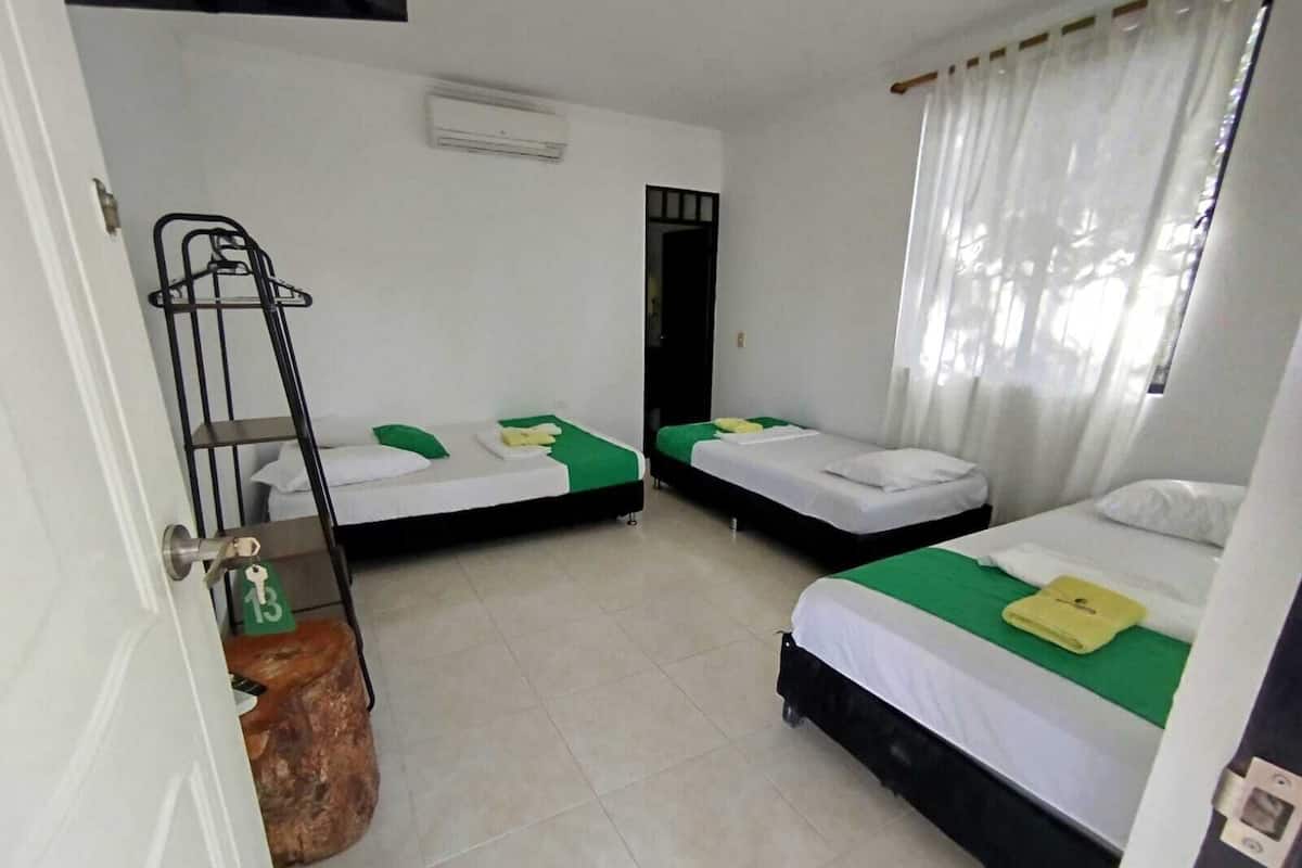 Habitació amb tres llits individuals, cadascun amb una colcha verd i blanca, coixins blancs i una tovallola groga. Hi ha una escala metàl·lica negra adossada a la paret, un aparell de aire condicionat blanc i una porta blanca amb un pany.