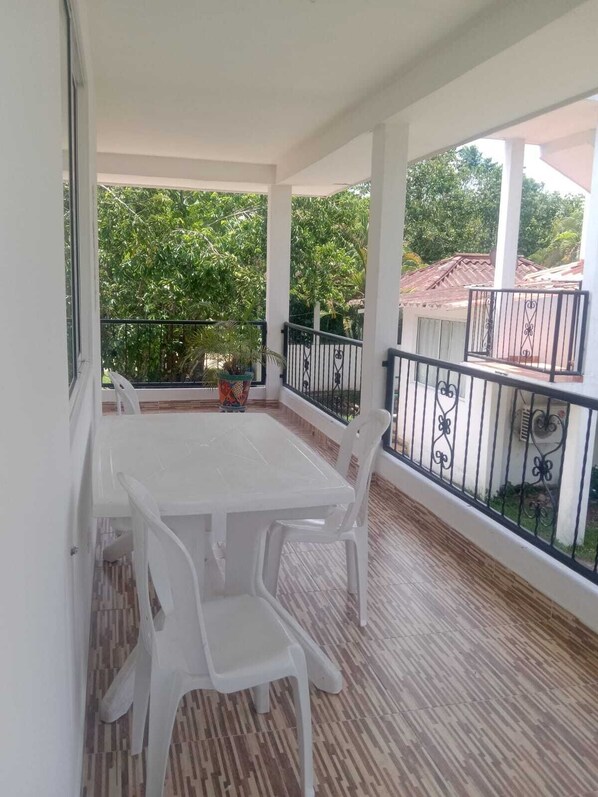 A covered balcony with a white table and chairs, a potted plant, and a view of greenery and buildings in the background. - Hotel Campestre Mastranto (Villavicencio)