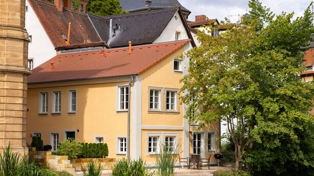 Un bâtiment jaune avec un toit rouge, un étang devant et un arbre sur la droite.