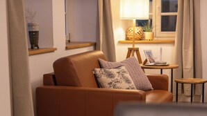 Un salon chaleureux avec un canapé marron, une petite table ronde avec une lampe et une fenêtre avec des rideaux.