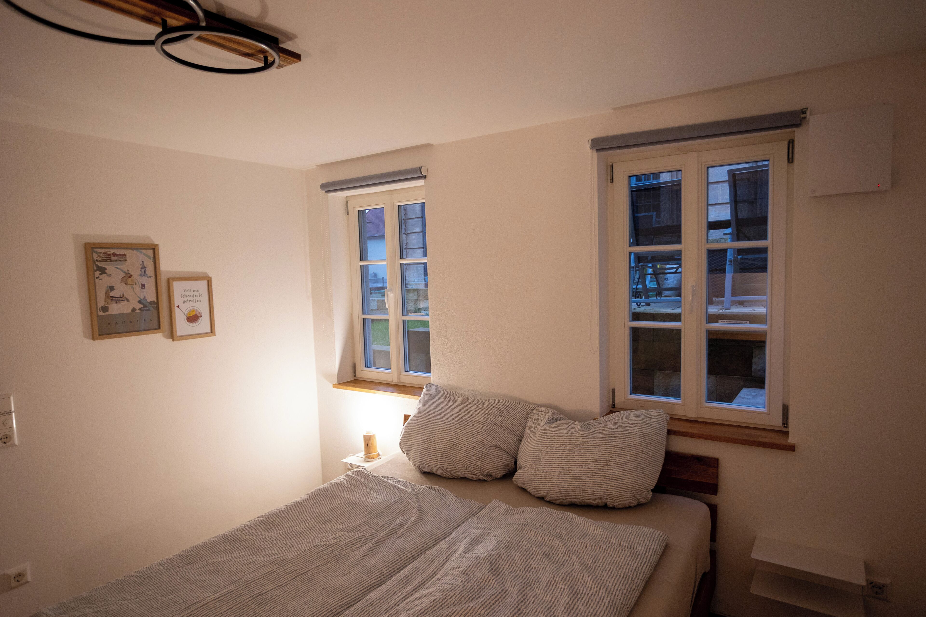 Ein Schlafzimmer mit einem Bett, zwei Fenstern, einem Nachttisch und einem Wandbild.
