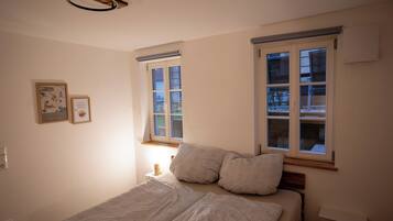 Ein Schlafzimmer mit einem Bett, zwei Fenstern, einem Nachttisch und einem Wandbild.