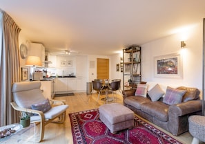 Uma sala de estar moderna com sofá, pufê e poltrona, uma área de jantar com mesa e cadeiras, e uma cozinha ao fundo.