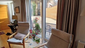 A room with two chairs, a table with a vase of flowers, and a view of a courtyard with a wooden fence and plants.