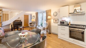A modern living room with a glass dining table, a sofa, and a kitchen area with white cabinets and a stove.