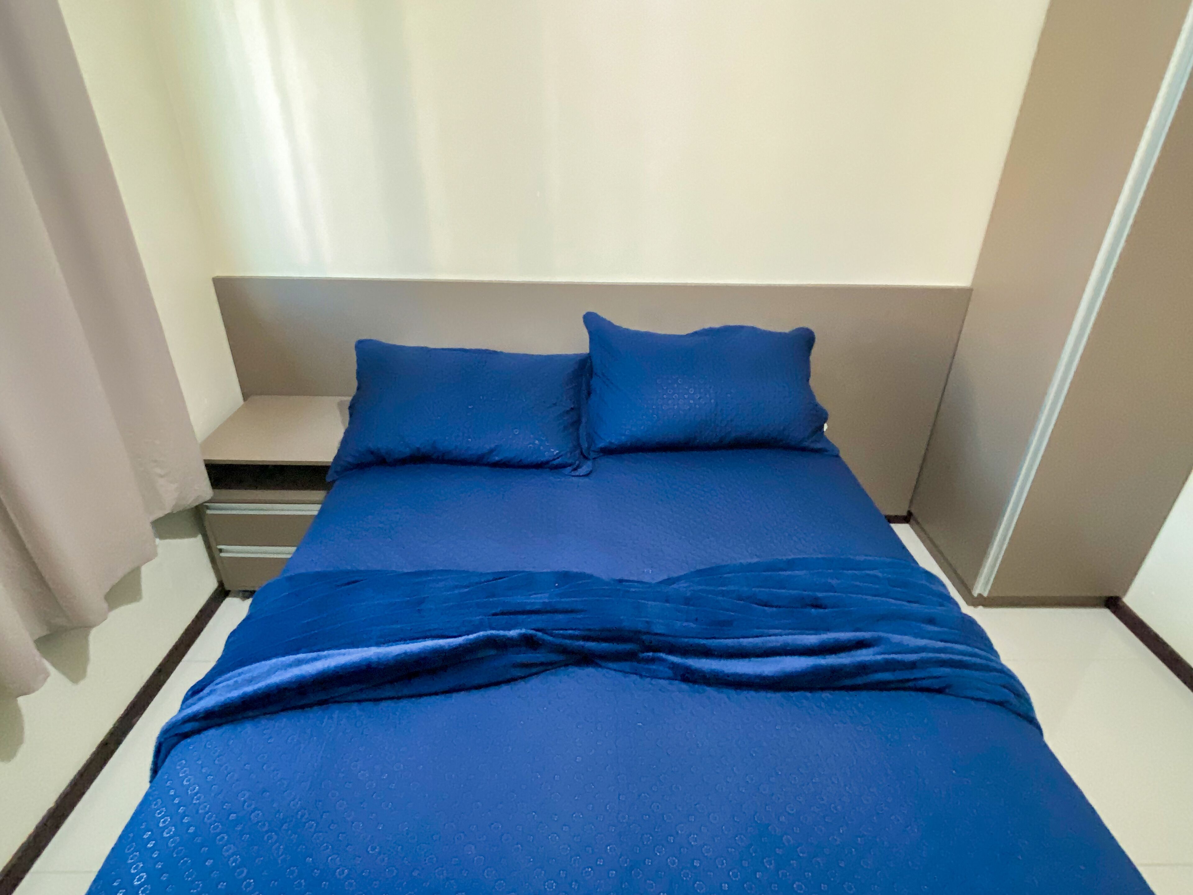 A bed with a blue bedspread and pillows, a bedside table, and a wall-mounted shelf.