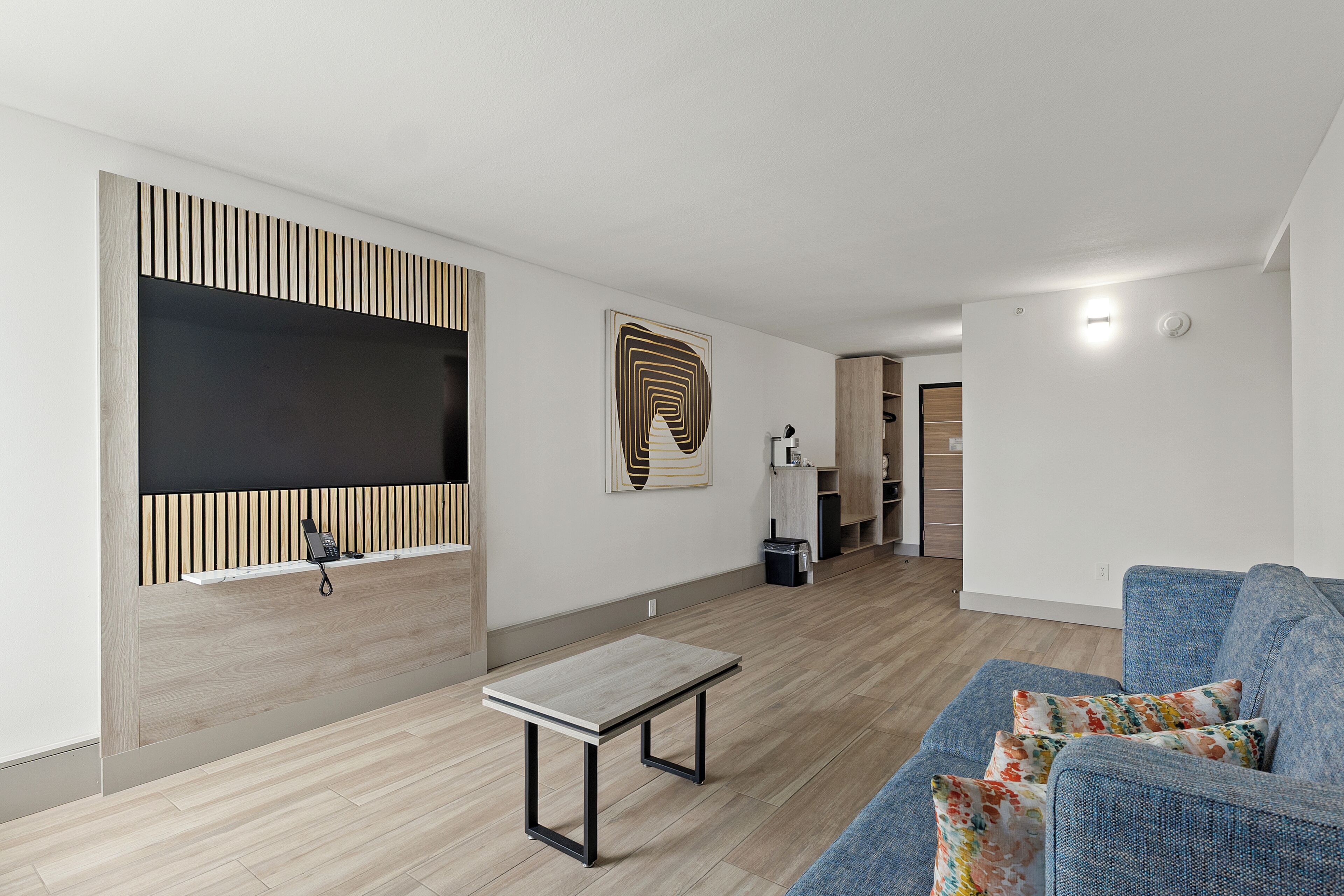 A modern living room with a sofa, a coffee table, a TV mounted on a wooden panel, and a patterned artwork on the wall.
