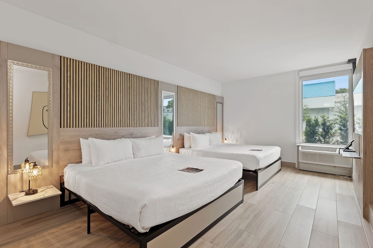 A hotel room with two beds, a large mirror, a wall-mounted light fixture, and a window with a view of buildings.