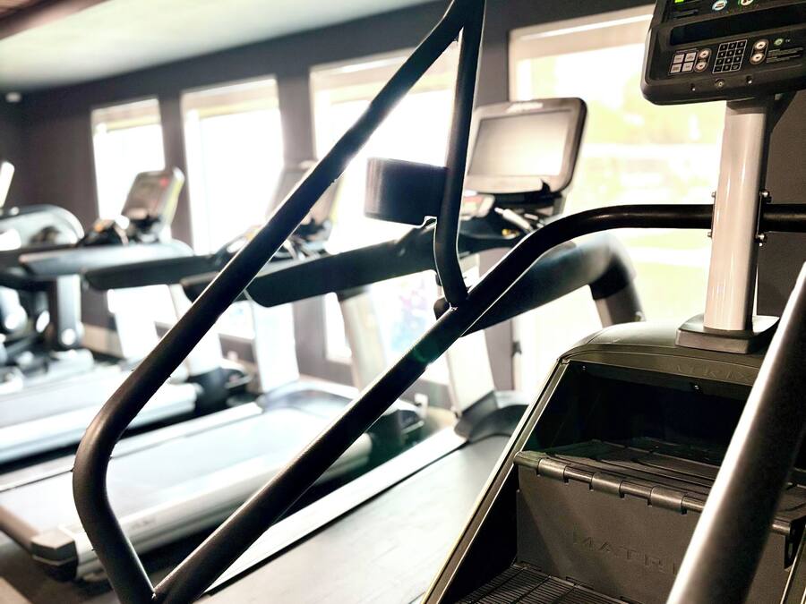 A treadmill with a digital display and a control panel, in a gym with exercise equipment and windows.