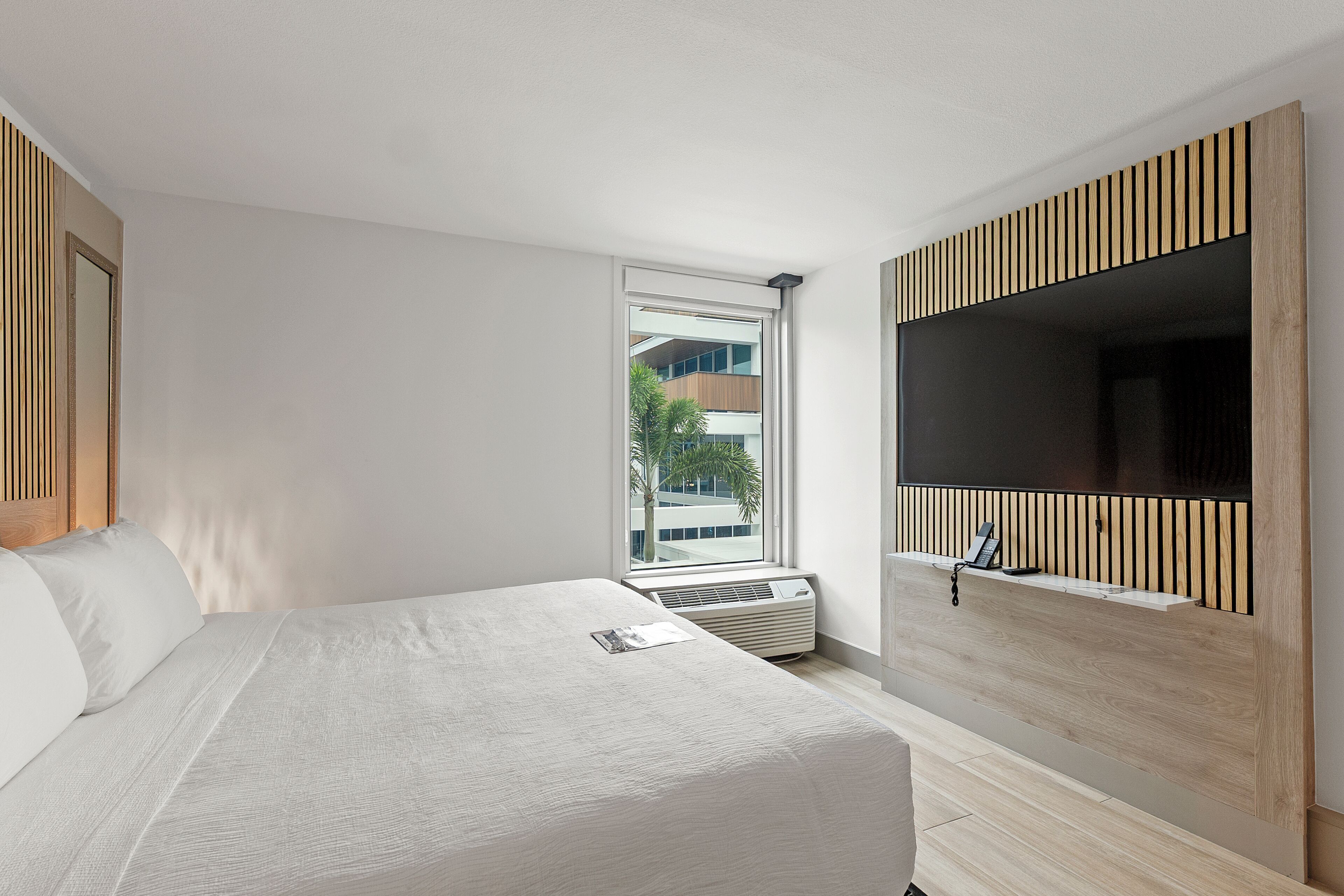 A modern hotel room with a bed, a large window, a flat-screen TV mounted on a wooden panel, and a built-in air conditioning unit.