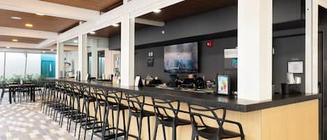 A modern bar area with a long counter and multiple stools, a flat-screen TV mounted on the wall, and a tiled floor.