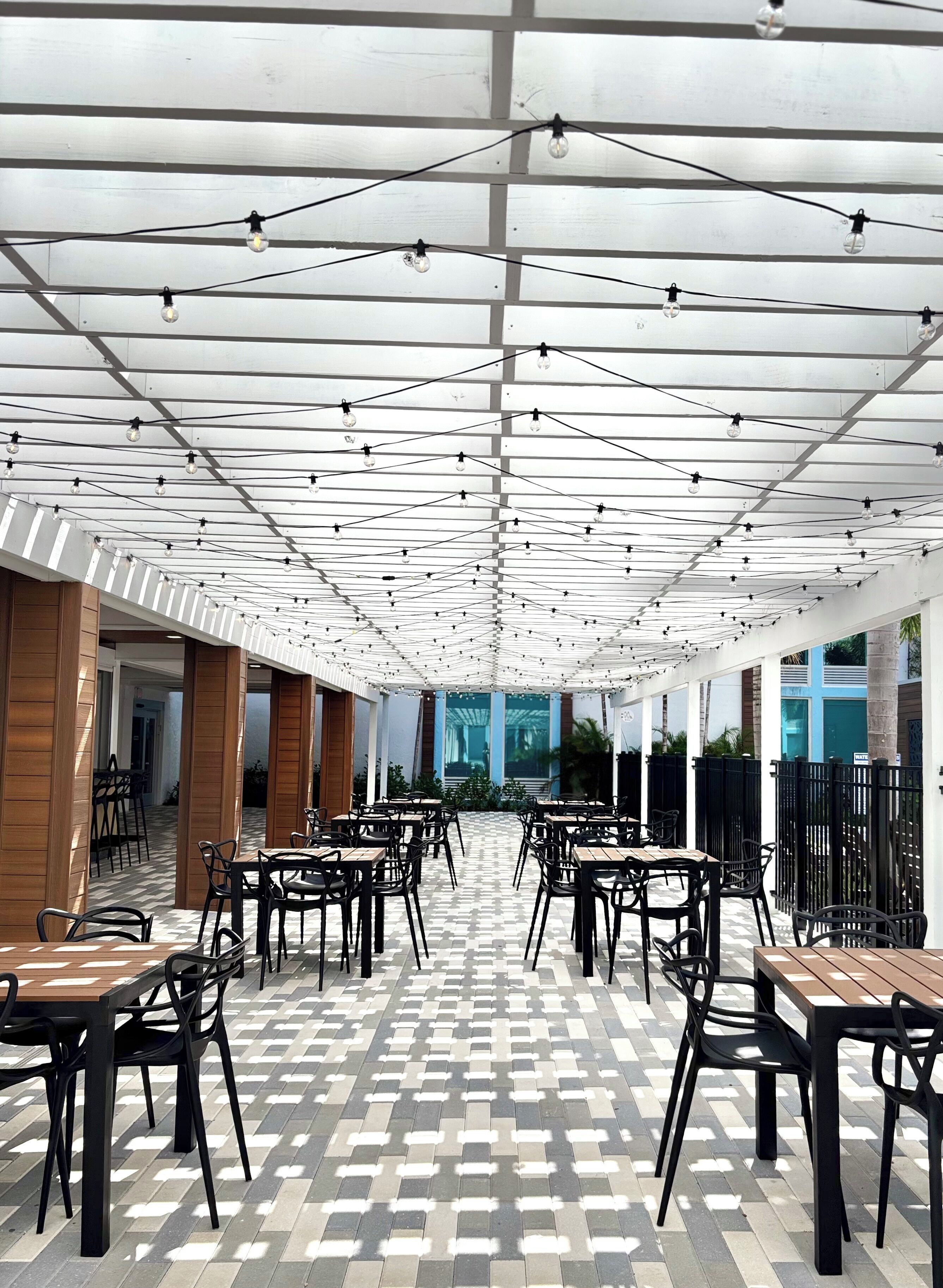 Un espace extérieur abrité, aménagé avec des tables et des chaises, doté d’un sol à motifs et illuminé par des guirlandes lumineuses suspendues au plafond.