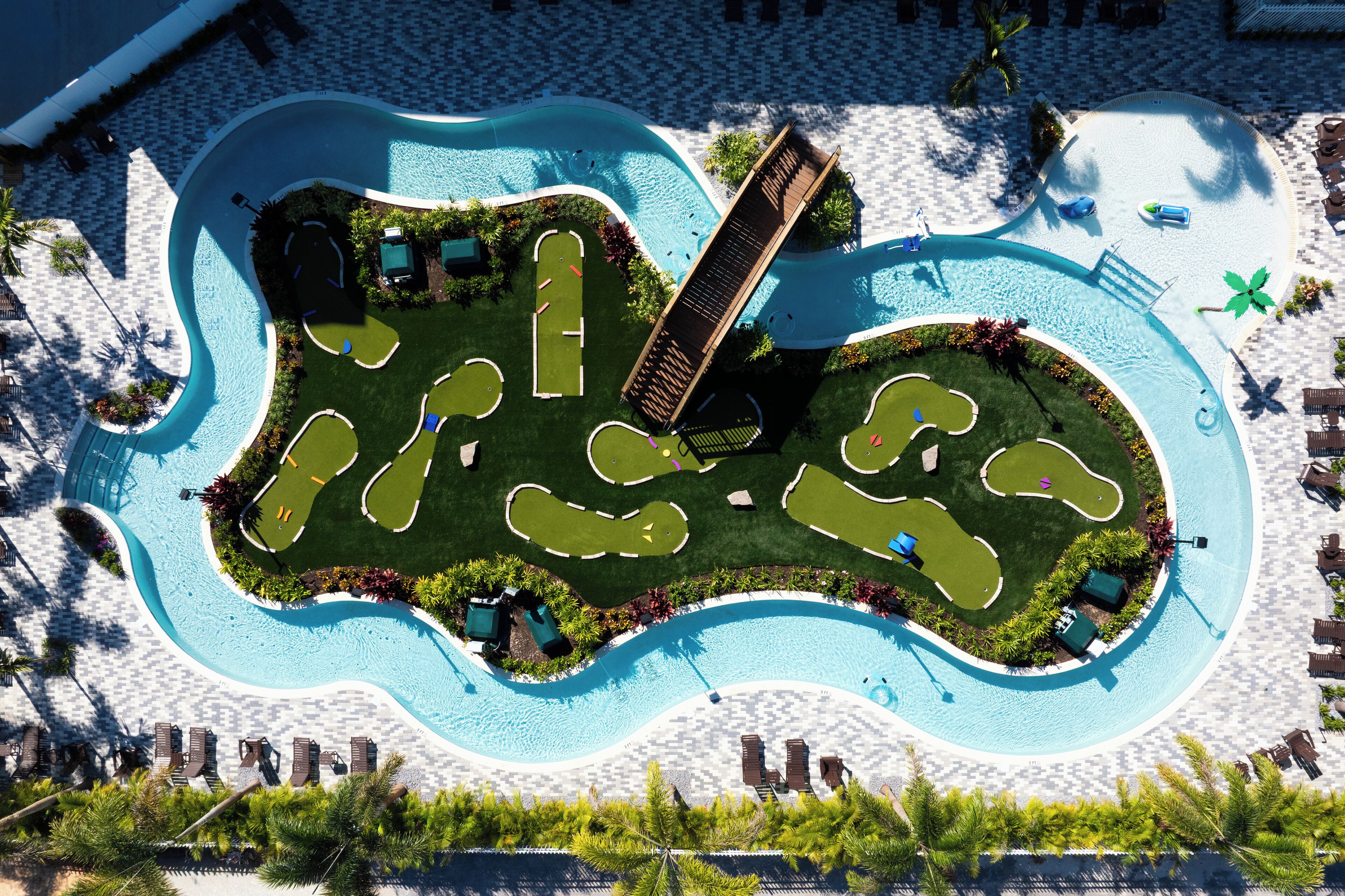 An aerial view of a resort with a large swimming pool, multiple lounge areas, and a well-maintained lawn.