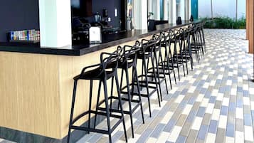 Un área de bar moderna con una barra alargada y varios taburetes, que presenta un patrón de piso a cuadros y detalles de techo de madera.