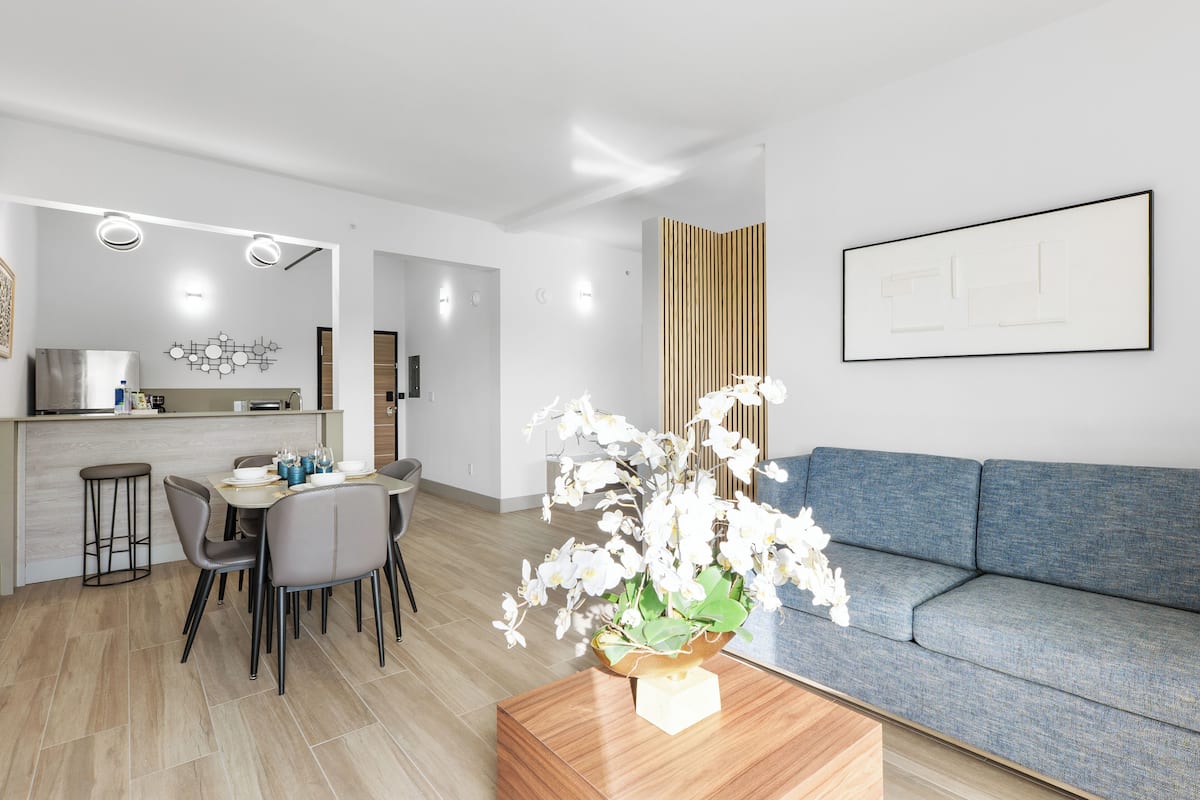 A modern living room with a blue sofa, a dining area with a table and chairs, and a kitchen in the background.