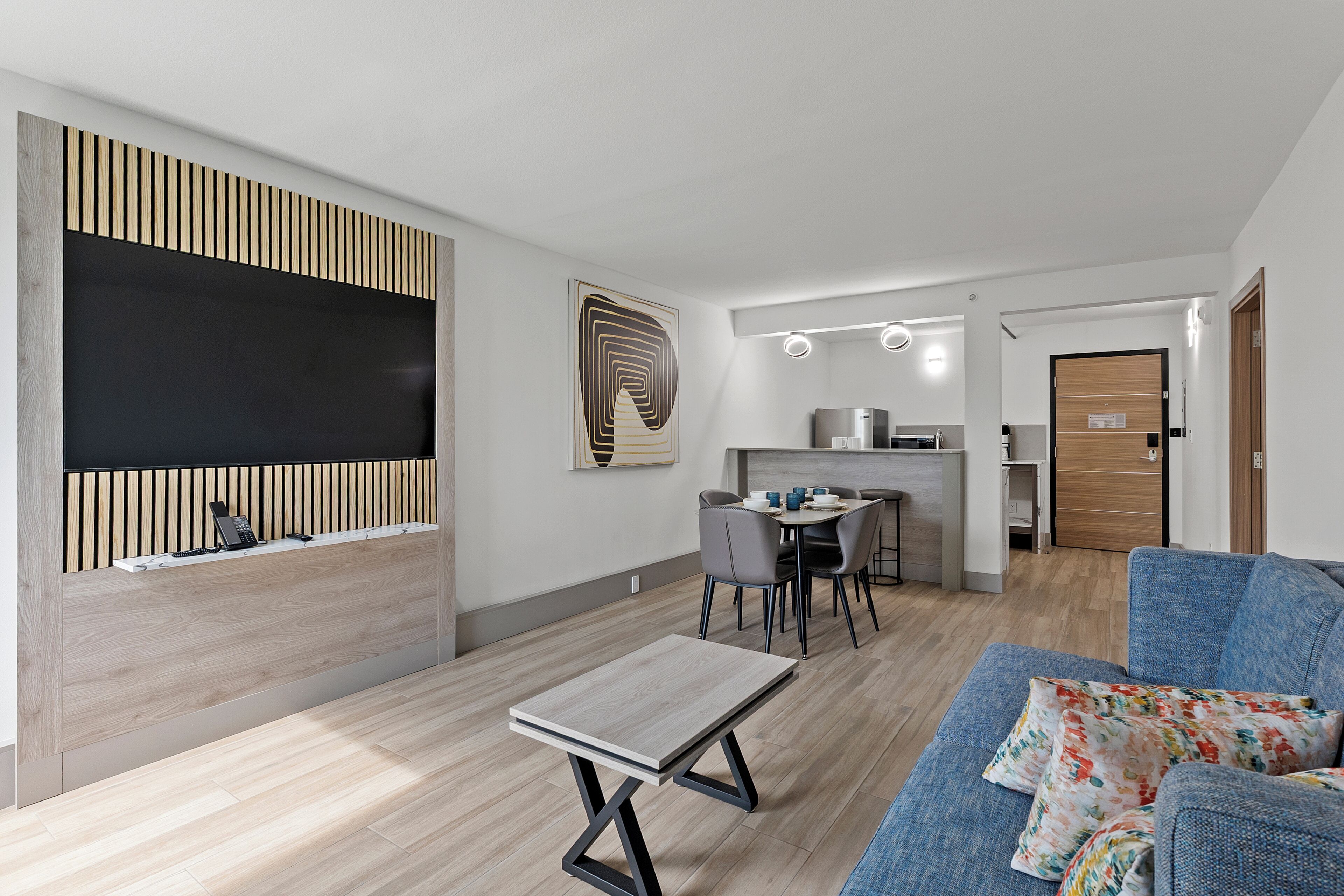 A modern living room with a sofa, a coffee table, a dining area with chairs, a kitchen in the background, and a large artwork on the wall.