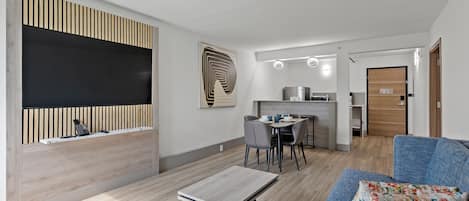 A modern living room with a sofa, a coffee table, a dining area with chairs, a kitchen in the background, and a large artwork on the wall.