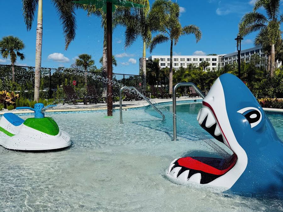 Zona de piscina con una fuente en forma de tiburón, palmeras y un edificio al fondo.