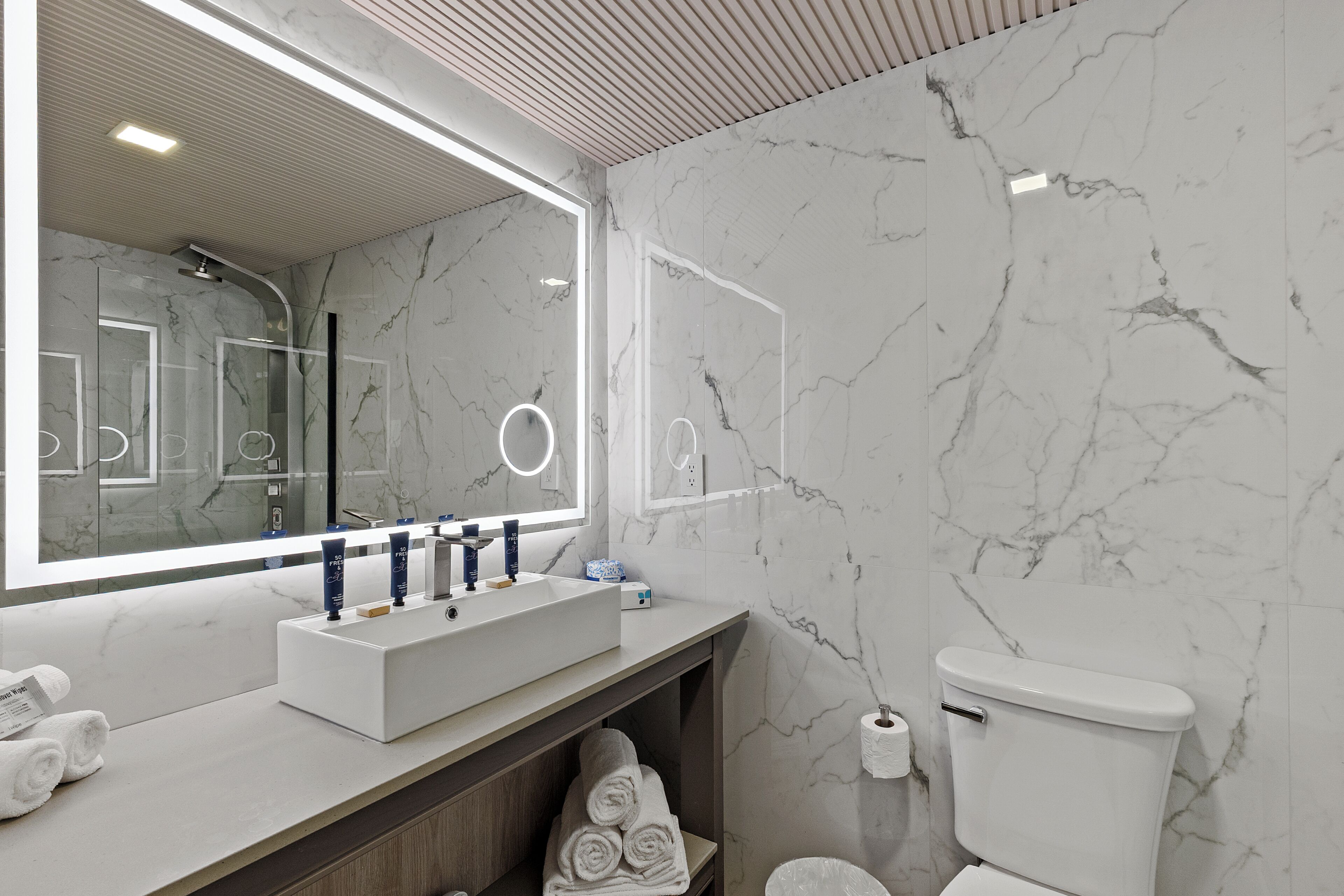 Ein modernes Badezimmer mit Marmor-Spritzschutz, einem großen Spiegel mit integrierter Beleuchtung, einem weißen Waschbecken inklusive Waschtisch und einer Toilette.