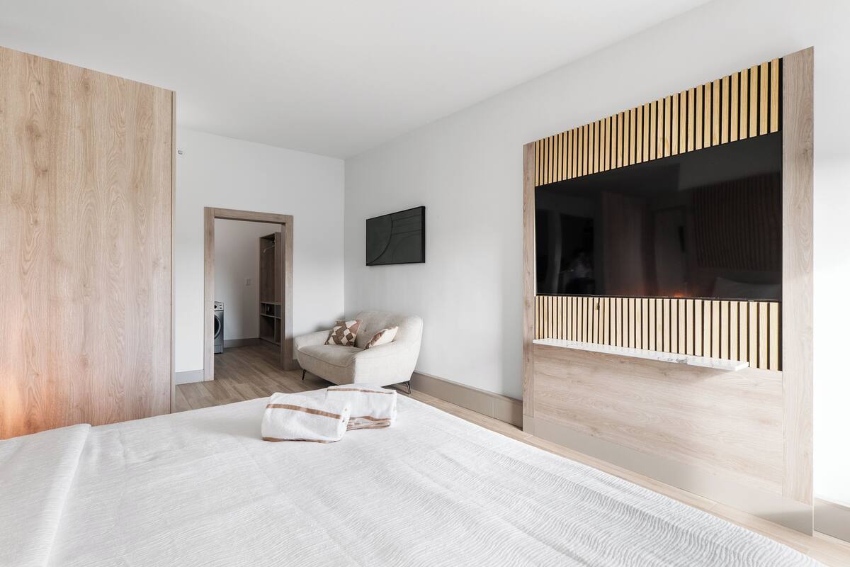 A modern bedroom with a large bed, a wooden accent wall, a TV mounted on the wall, and a chair with a cushion.