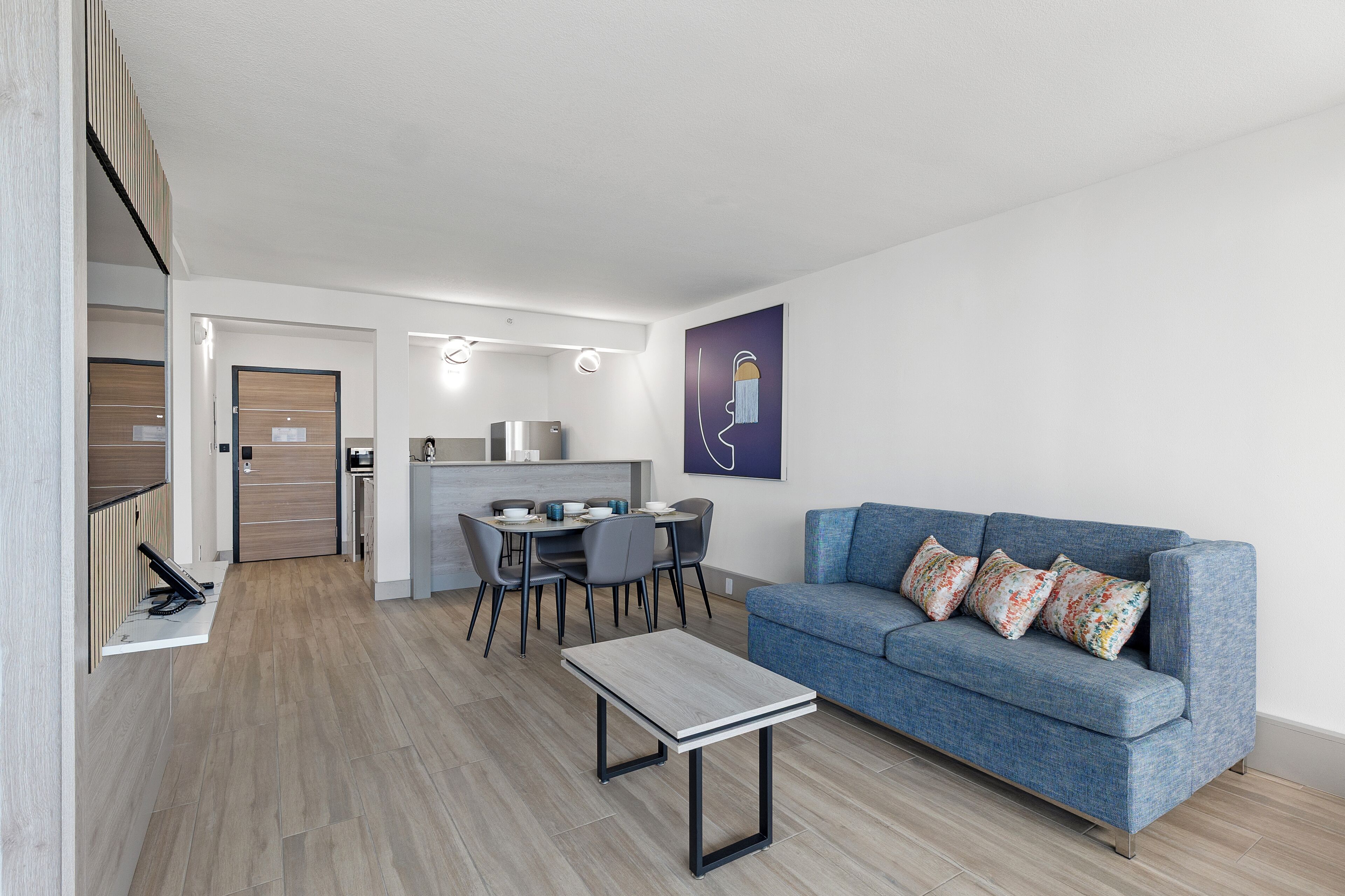 En moderne stue med en blå sofa, et lille bord og et spiseområde med stole, samt et køkken i baggrunden.