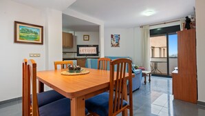 A dining area with a wooden table and blue upholstered chairs, a kitchen in the background, and a view through a glass door. - Apartment "Deltebre Riu" with Shared Pool & Wi-Fi (Deltebre)