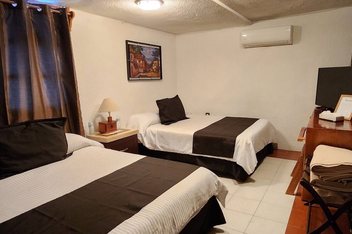 A hotel room with two beds, a desk, a chair, an air conditioner, and a painting on the wall.