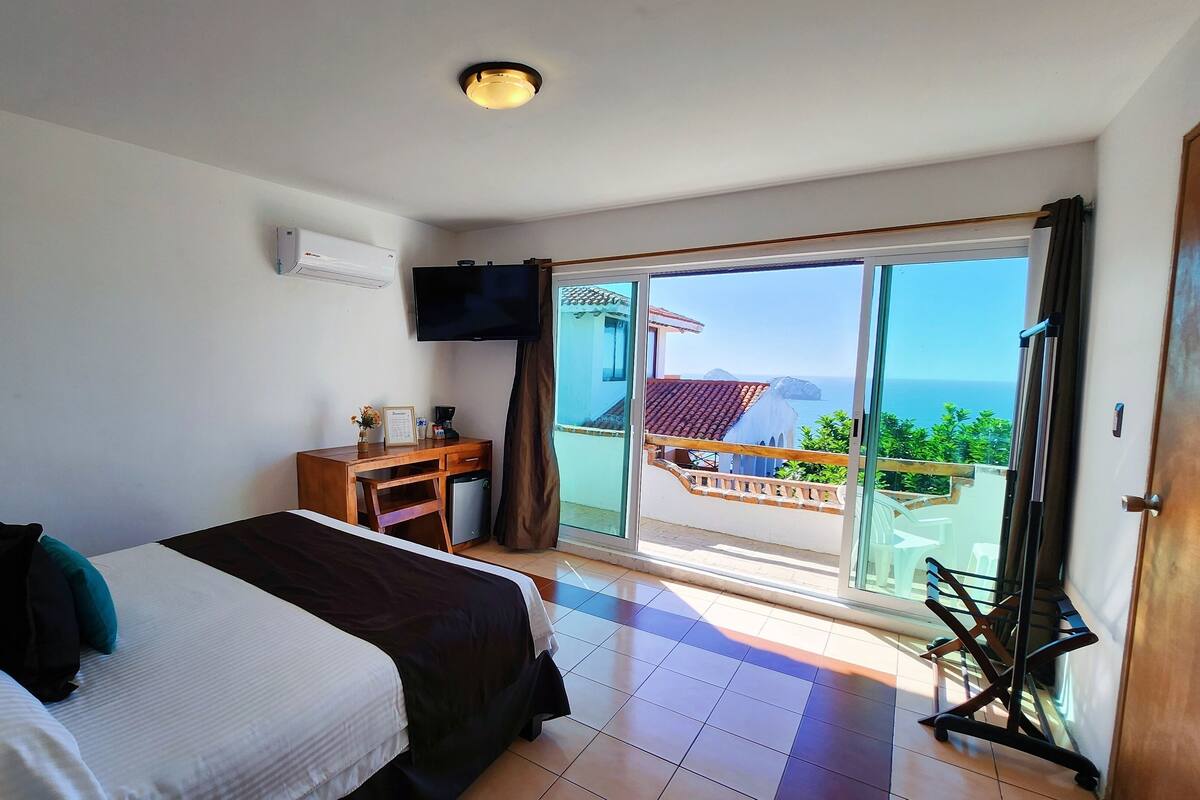 A hotel room with a bed, a desk, a television, and a balcony with a view of the sea.