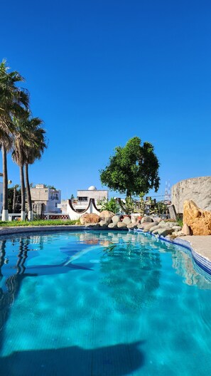 Une piscine d’un bleu éclatant, bordée de carreaux, entourée de rochers et de palmiers, avec des bâtiments visibles au loin.
