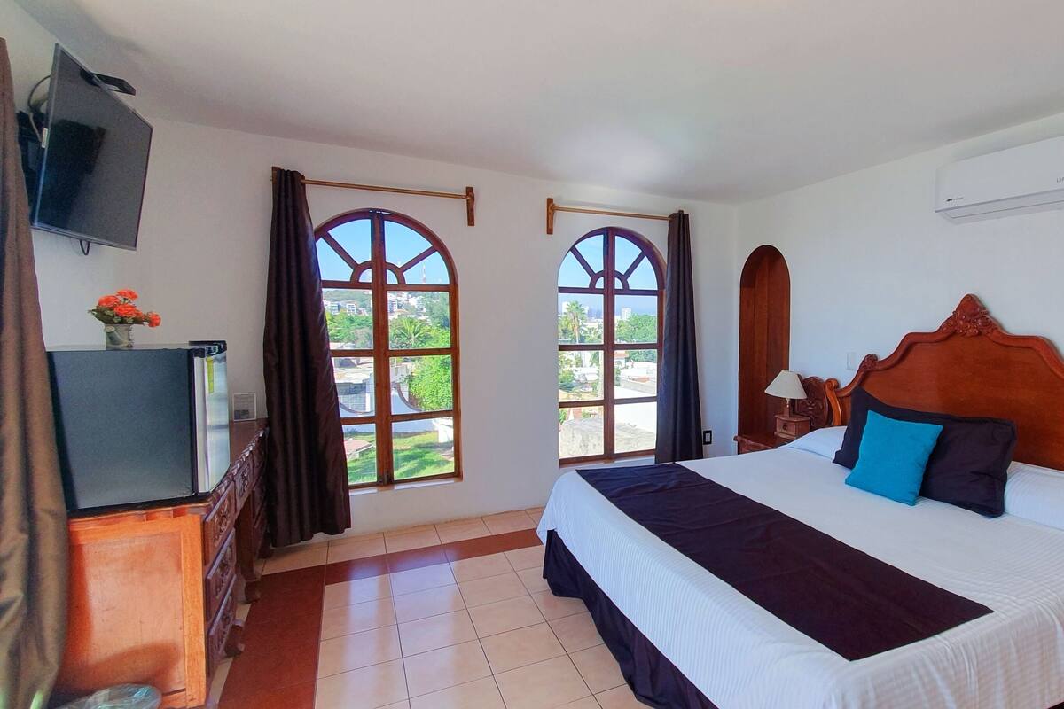 A hotel room with a large bed, a TV, and two windows with curtains.