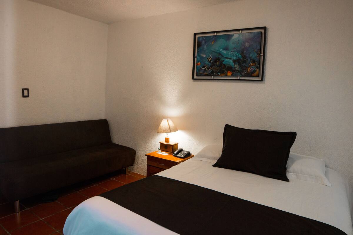 A hotel room with a bed, a nightstand with a lamp, a couch, and a painting on the wall.