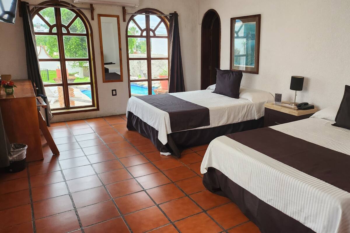 A hotel room with two beds, a desk, a chair, a trash bin, and large windows with a view of a swimming pool.