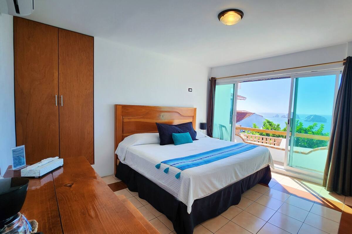 A bedroom with a bed, a wooden wardrobe, a desk with a computer, and a balcony with a view.