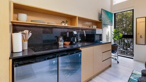 A modern kitchen with a black countertop, wooden cabinets, and a dishwasher. There are shelves with dishes and a potted plant. - Olive Apartments (Miami)
