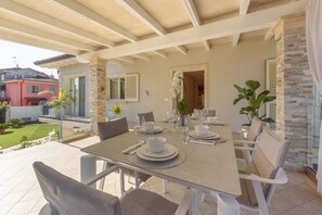 Un espace extérieur aménagé pour les repas, avec une table dressée pour quatre personnes, entourée de chaises, et offrant une vue sur un jardin et un bâtiment en arrière-plan.