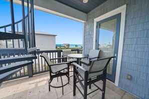 Een overdekt terras met een tafel en stoelen, uitzicht op het strand, en een gebouw met een blauwe gevel.