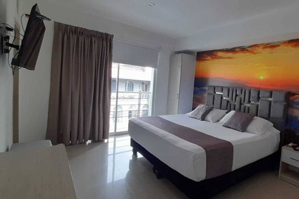 A hotel room with a large bed, a nightstand, a TV mounted on the wall, and a window with curtains.
