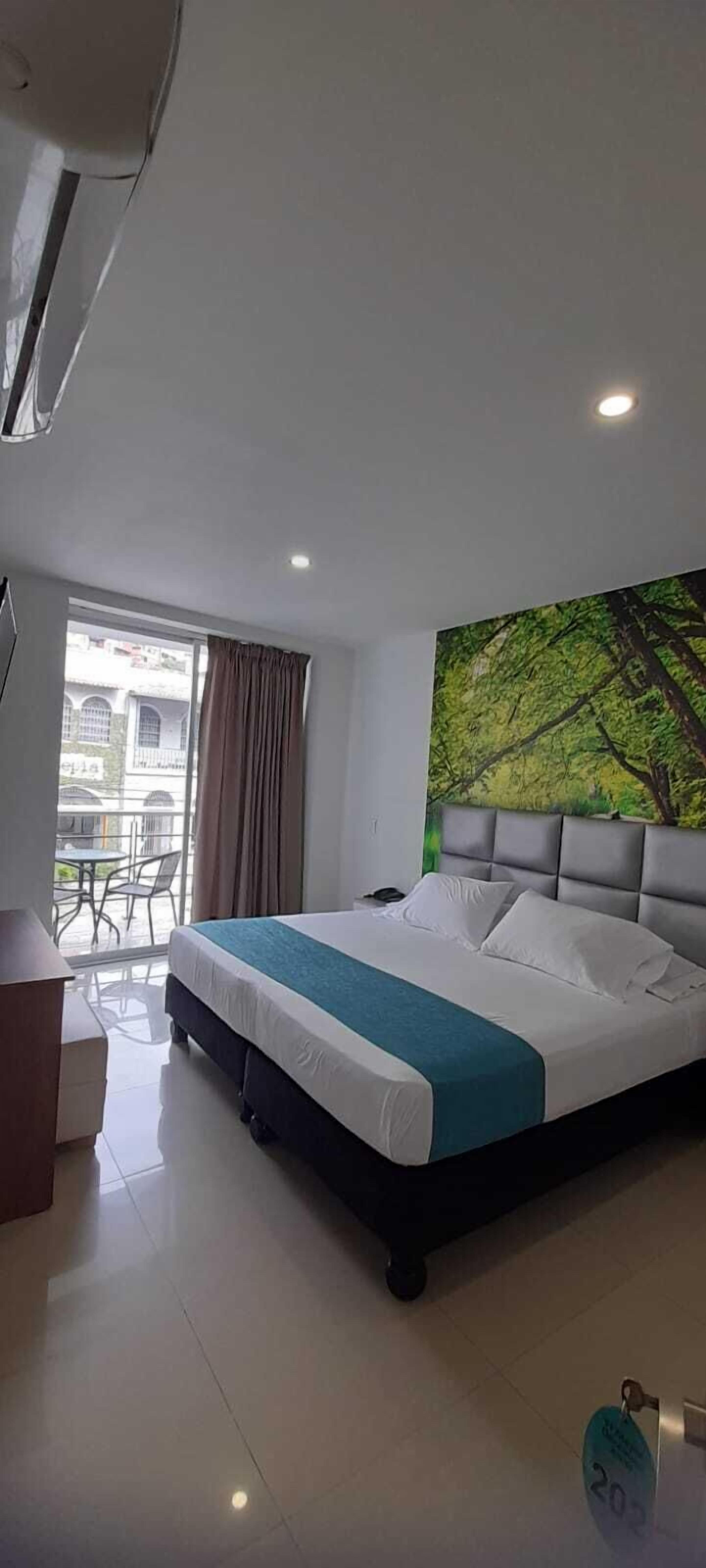 Habitación de hotel con cama, ventana con cortinas, un cuadro en la pared y un balcón con sillas.