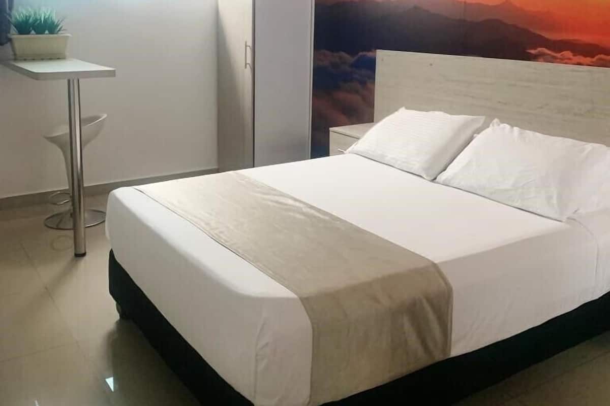 A neatly made bed with white linens in a room with a large painting of a sunset on the wall.