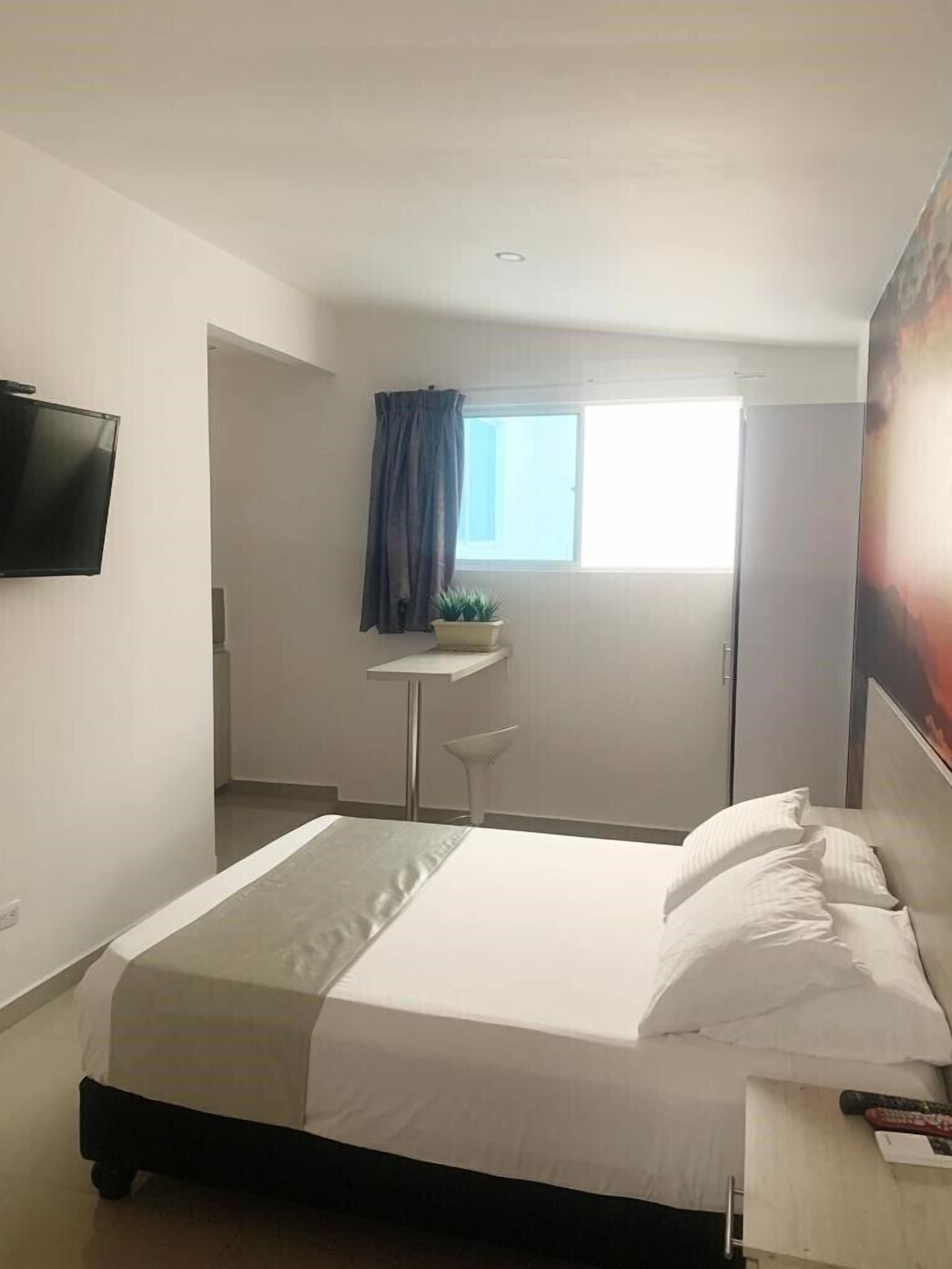 Habitación de hotel con cama, televisor montado en la pared, una mesita con una planta y una ventana con cortinas.