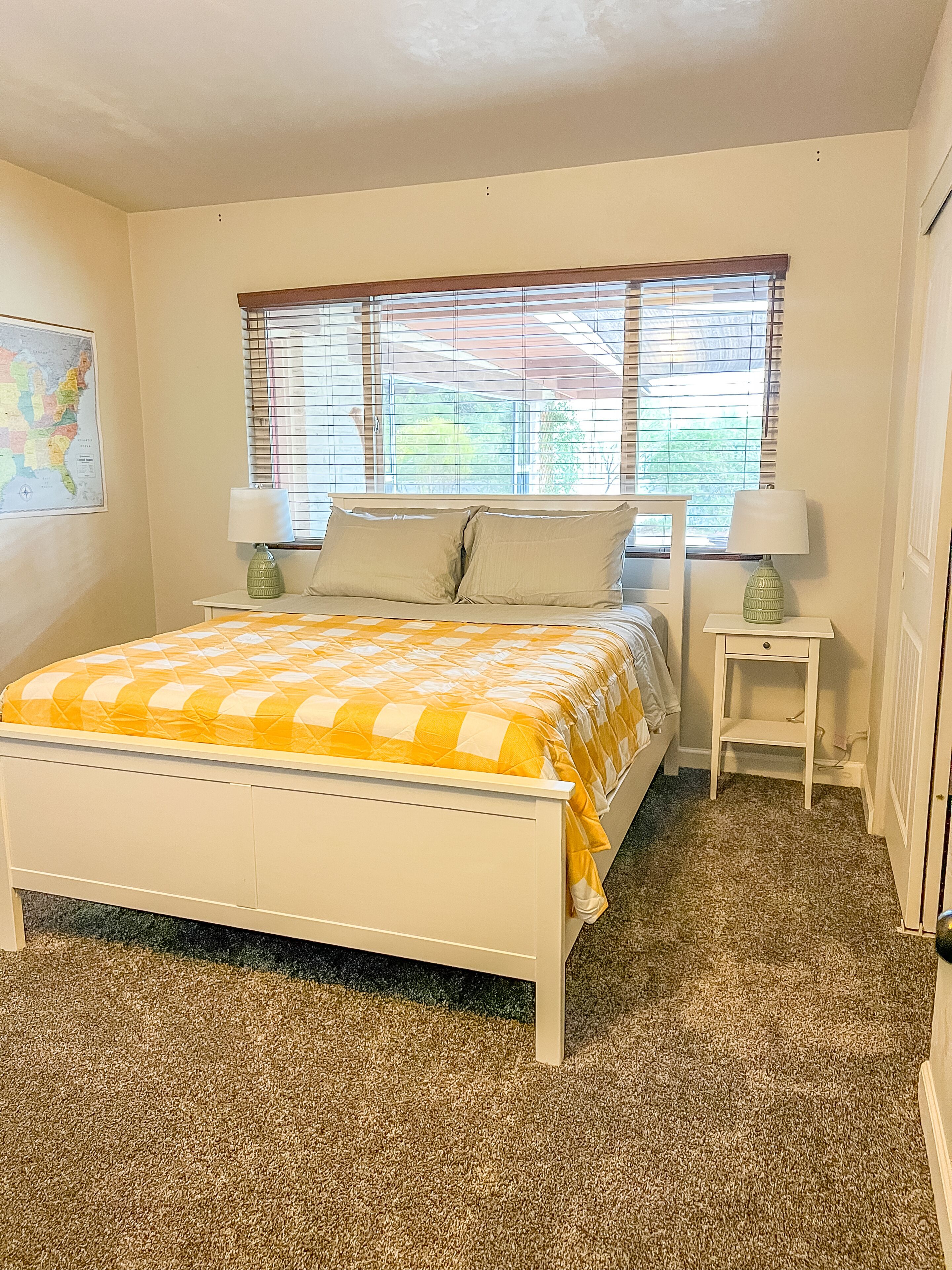 A bedroom with a bed, two bedside tables, a window with blinds, and a map on the wall.