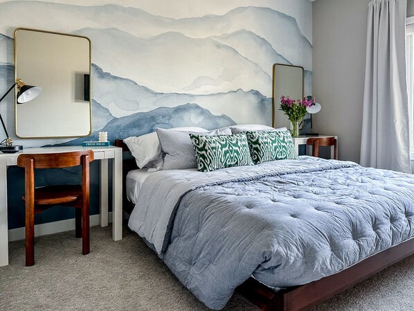 A bedroom with a bed, a desk, a chair, and a wall with a mountain painting.