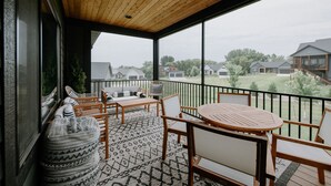 Un balcón techado con muebles de madera, un tapete estampado y vista a casas al fondo.