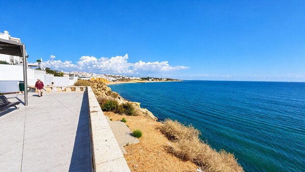 A coastal walkway with a clear view of the sea, a paved path, and a person walking.